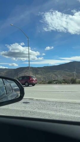 The video shows a driver's perspective of driving on a highway on a bright, sunny day. The sky is blue with scattered white clouds, and there are mountains in the background. Several vehicles, including trucks and cars, can be seen on the road. The camera is inside the car, focusing on the road ahead, and occasionally showing a side mirror with a reflection of the sky and other vehicles. A sign for "Exit 104" is visible, as well as a billboard advertising "Best Spa." Upbeat music plays in the background.