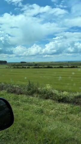 The video is filmed from inside a vehicle looking out through the windshield. The windshield has a few water droplets or smudges on it. Outside, there is a green field of grass that extends to the horizon. In the distance, there are some trees and buildings. The sky is blue with fluffy white clouds. The side mirror of the vehicle is visible in the lower left corner of the frame. The scene appears to be a rural landscape on a cloudy day.
