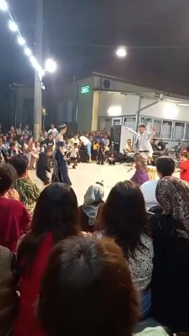 The video captures an outdoor gathering at night. Several people are present, with a group seated in the foreground facing a central area where a performance is taking place. In the center, a woman in a long blue dress stands near a man in a white suit who appears to be directing or performing. Several children are also in the performance area, some kneeling or dancing. Other individuals, some holding what might be instruments or cameras, are positioned around the performers. A string of lights illuminates the scene, suggesting a festive or celebratory atmosphere. The background shows buildings and trees, indicating an outdoor venue. The audience in the foreground is varied in age and attire, with many women wearing headscarves. The overall impression is that of a lively community event or performance.