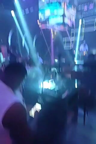 The video shows the interior of a nightclub. People are seen socializing and dancing in a dimly lit environment with flashing lights. A DJ booth is visible in the background, with a DJ playing music. There are tables and seating areas, as well as a large screen displaying abstract visuals. The overall atmosphere appears to be lively and energetic.