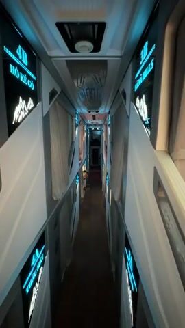 A first-person perspective video shows the interior of a bus designed with sleeping compartments. The camera moves down a narrow aisle, with sleeping berths visible on both sides. Each berth appears to have a curtain for privacy. Neon blue lights illuminate the signs above the berths, which display numbers such as "4B" and "3B." Some berths have small decorative images of mountains. The ceiling of the bus is visible, with ventilation systems and lighting fixtures. A person is partially visible peeking out from one of the berths. Towards the end of the video, the camera pans to the right, revealing a more detailed view of one of the sleeping compartments, which includes a bed with bedding and an orange accent. A small fan is mounted on the wall of this compartment, and a green display screen is visible above the bed. The overall impression is a modern, somewhat futuristic design for overnight bus travel.
