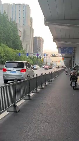 The video shows a sidewalk with a metal guardrail next to a multilane road. The road is filled with cars, buses, and motorcycles moving in both directions. A row of traffic lights hangs overhead. High-rise buildings and some trees line the side of the road. The sky is a hazy orange color, indicating it's dusk or dawn. The camera is stationary.