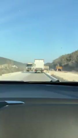 The view from inside a car driving on a road approaching road construction is captured in this video. In front of the car, there are two trucks, one a dump truck and another a white transport truck with the number "3" in red on the back. To the right of the road, an excavator is visible. The road is lined with bare trees and dusty terrain, with a clear blue sky overhead. Other vehicles can be seen moving in the distance amidst the dust. The perspective is from the driver's seat, focusing on the road ahead.