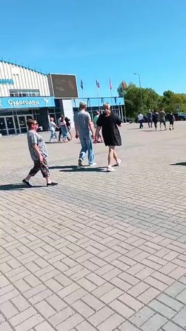 The video captures a sunny outdoor scene, likely a paved area or parking lot. Several people are visible, some walking towards the camera, others away, and some standing. In the background, there's a large building with a distinctive curved roof and blue lettering. Cars are parked in the distance, and trees line the horizon, suggesting a mix of urban and natural elements. The ground is paved with rectangular light grey blocks. The sky is clear and blue.