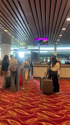 Passengers are queuing at an airport gate. Most have luggage with them. The gate agent is behind a counter. The overhead flight board shows flight OD 0171 to Denpasar and EY 5615 to Akron, among others. The carpet is red with a textured pattern. The ceiling is dark brown with horizontal beams.