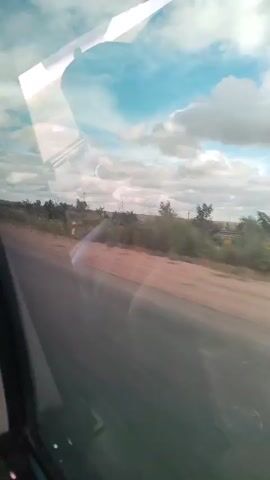 This video shows a view from inside a vehicle looking out the window. A road or dirt path is visible, with sparse vegetation and some trees lining the side. In the distance, a few wind turbines can be seen against a cloudy blue sky. The reflection of what appears to be the interior of the vehicle is visible on the window. The camera is moving, creating a sense of travel.