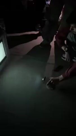 In a darkened space, two people, seen from their mid-torso down, are near a turned-on television set. The television screen displays Bulgarian text. One person, wearing a burgundy-colored top and a watch, holds a small object, possibly a lighter, and moves it near the floor as though inspecting it. The other person wears darker trousers and shoes. The shadows cast by the individuals indicate a light source is near them, also illuminating the floor and television set. A mobile phone is lying on the floor near the person holding the object. The overall environment appears to be dimly lit and possibly indoors.