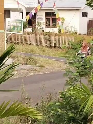 A woman wearing a hijab and a patterned shirt rides a black scooter down a paved road. She is wearing a yellow skirt. She is partially obscured by palm fronds and other green foliage. In the background, there is a white building with a fence and a sign. The road appears to be in a rural or suburban area.