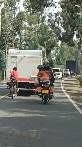 The video shows a road scene in what appears to be a rural or semi-rural area. A person on a bicycle wearing a red shirt is on the left side of the frame, riding close behind a white truck with an orange stripe along the bottom. The truck's license plate number is partially visible and reads "4196BL 01". To the right of the truck, there is a motorcycle with three people riding it, all wearing helmets. The person in the middle is wearing an orange safety jacket and pants. The motorcycle's license plate number is "RE 429F". The road has a yellow dividing line, indicating two lanes. The road is lined with tall trees on both sides. In the distance, more vehicles are visible, including vans and trucks. The sky is overcast, casting soft light on the scene.