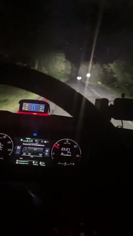 A nighttime first-person view from the driver's seat shows a car moving along a dark, winding road, illuminated by its headlights. The dashboard is visible with lit instrument panels displaying information such as speed, time (23:27), and temperature (14°C). A small digital display above the instrument panel shows tire pressure readings. Trees line the road on either side, and two indistinct streetlights are visible ahead.