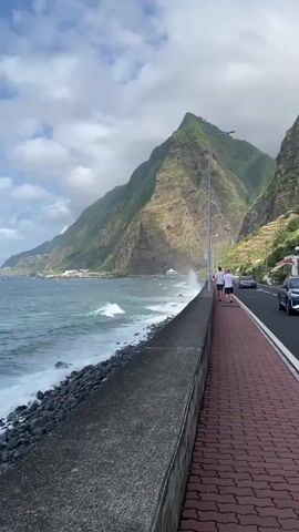 The video shows a scenic coastal road with a paved pedestrian walkway. Two people are walking on the walkway away from the camera. To the left of the walkway is a stone barrier, and beyond that, the ocean with waves crashing against a rocky shore. In the background, a steep, green mountain rises dramatically, with some buildings and terraced fields visible on its slopes. A road runs parallel to the walkway, and a car is visible driving on it. The sky is partly cloudy, with patches of blue visible.