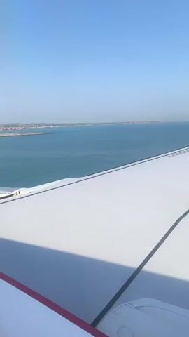 This video shows a view from an airplane window as it descends towards land. The camera is focused on the white airplane wing in the foreground, with a thin red stripe along its edge. Beyond the wing, a vast expanse of turquoise water stretches out, meeting a distant coastline dotted with buildings and vegetation. The sky is a clear, brilliant blue. The plane appears to be approaching land over the water, and the narrator states that they will be landing soon. The overall impression is one of arrival and the anticipation of reaching a destination.