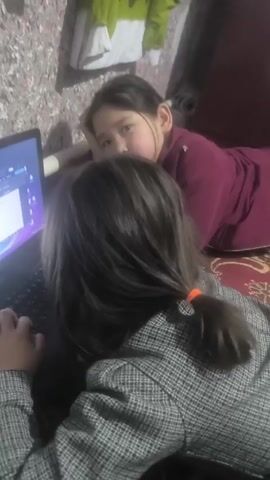 Two young girls are looking at a laptop screen. One girl is lying down, resting her head on her hand while gazing at the laptop screen. She is wearing a dark burgundy sweater. The second girl is sitting and typing on the keyboard of the laptop. She has her hair in a ponytail with an orange hair tie and is wearing a grey and black plaid jacket. The laptop is on a rug. The background includes a floral wallpaper and some clothing hanging in the upper left corner. Someone, probably an adult, is off camera speaking to the girls.
