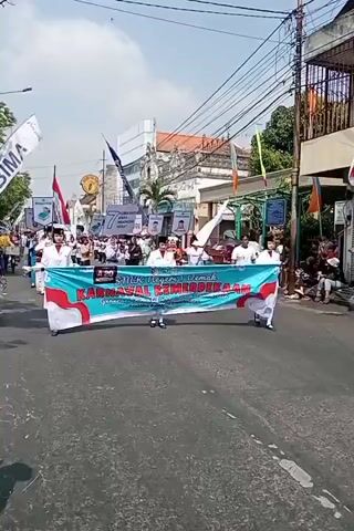 This video shows a parade in a street under a bright sky. A group of students, mostly wearing white uniforms with red trim and some wearing black hats, are marching forward. They are holding a large banner that reads "SMK NEGERI 1 DEMAK KARNAVAL KEMERDEKAAN". The banner is teal and red and has text in white. Several other banners and flags are visible in the background, along with buildings lining the street and power lines overhead. Some spectators are visible on the side of the road. The students in the foreground are holding the banner horizontally, with four students positioned at the corners and two in the middle. They appear to be walking in unison. The overall atmosphere seems festive.