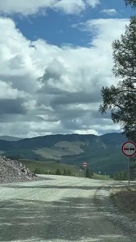 A POV shot from a vehicle driving on a gravel road reveals a vast landscape of rolling green hills and mountains under a partly cloudy blue sky. A traffic sign with a red circle and a horizontal bar indicating "no entry" is visible on the right side of the road. The road curves to the right, leading into the expansive scenery. The edges of the frame are bordered by trees on the right and a pile of rocks on the left.
