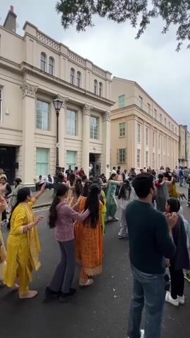 The video captures a street scene in front of a light-colored, classical-style building with columns. A group of people, primarily women and children, are dancing in a circle in the street. Many are wearing traditional Indian clothing, including brightly colored saris and salwar kameez. Some individuals are holding hands as they move in a circular motion. The atmosphere appears festive. The ground is paved, and there are trees with green foliage visible at the top of the frame. The sky is overcast. The perspective is from behind the dancing group, looking towards the building.