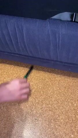 The video shows a close-up shot of a blue sofa with a textured cork floor. A hand enters the frame from the left, holding a green and black object, possibly a tool or pen. The hand moves the object underneath the sofa. The rest of the video continues to show the hand and the object moving beneath the sofa. The lighting suggests an indoor environment.