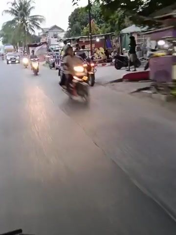 The video captures a street scene, possibly in a developing country, showing various vehicles and people. In the foreground, a person's finger is visible, suggesting the recording is from a handheld device. The road is paved and appears to be a public thoroughfare.

At 00:00, a three-wheeled vehicle, likely a tuk-tuk or auto-rickshaw, with passengers in the back, is moving away from the camera. Other motorcycles are also visible on the road.

At 00:01, a motorcycle with its headlights on is moving towards the camera. The background shows buildings and trees.

At 00:02, a dark-colored car with its headlights on is approaching. The car has its lights on, indicating it might be dusk or a dimly lit area.

At 00:03, a person is riding a motorcycle towards the camera. A yellow truck is visible further down the road, also moving away from the camera.

At 00:04, the scene is blurred, but the yellow truck and motorcycles are still discernible.

The overall environment appears to be a town or city street with commercial establishments and residential buildings lining the road. The time of day seems to be either early morning or late evening, given the lighting conditions and the use of headlights by some vehicles. The activity on the street suggests it is a busy thoroughfare.