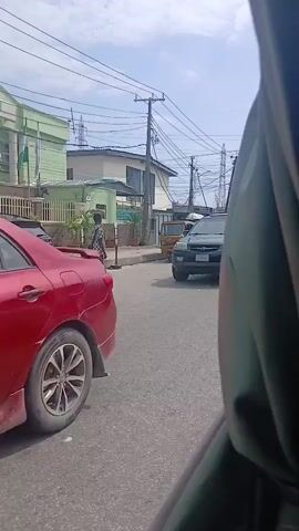 A black SUV drives past a black sedan. A person stands on the side of the road, looking in the direction of the cars. In the background, a green building with flags and a fence is visible. A tuk-tuk is also seen further down the road, along with other cars. Overhead power lines crisscross the sky, which is partly cloudy. The video is taken from inside a vehicle, with the side of the vehicle visible in the foreground.