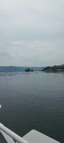 A calm, grey, overcast day with a wide expanse of rippling water. In the distance, a small, green, tree-covered island sits to the left, with some structures on it and connected to the shore by a bridge. To the right, a shoreline is lined with houses built on stilts over the water. In the middle of the water, a person in a small boat is moving away from the camera. The boat appears to be a narrow canoe or kayak, and the person is facing away from the camera.