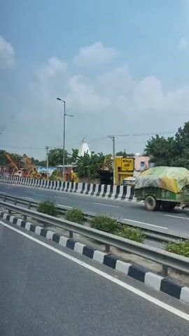 The video shows a highway scene with construction equipment, including cranes, and a small white temple-like structure with a white dome visible in the background. The road is paved with a metal barrier separating it from the area where the construction and temple are located. The sky is overcast with scattered clouds. The perspective suggests the video is being recorded from a moving vehicle.
