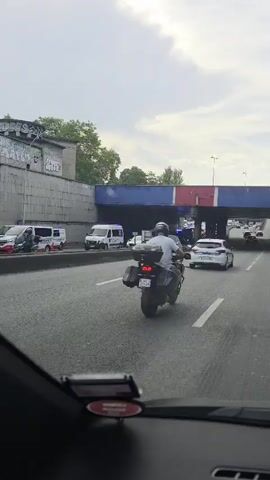 The video shows a view from inside a car driving on a multi-lane highway. The weather appears overcast, with a cloudy sky. The setting is urban, with a large concrete overpass with red, white, and blue painted stripes spanning the highway. There is graffiti on a concrete wall to the left of the highway.

Several vehicles are visible on the highway, including vans, cars, and a motorcycle. Some vehicles appear to be emergency services, indicated by flashing blue lights, possibly police or ambulance. The traffic appears to be moving slowly, suggesting a traffic jam or an incident ahead. The time of day seems to be daytime, likely late morning or afternoon, due to the ambient light.

Notable details include a motorcycle with a rider wearing a helmet and a white top, with saddlebags on the back. A black hatchback car is prominently featured in the foreground to the left, with a license plate that reads "BG 308 SW". Further ahead, a white car and other vehicles are visible. The overall atmosphere suggests a moment of travel through a busy urban environment with potential disruption. No discernible emotions are conveyed beyond the factual observation of traffic and vehicles.
