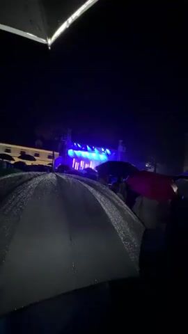A nighttime outdoor concert scene is depicted. The foreground is dominated by several open umbrellas, some glistening with water, suggesting rain. Beyond the umbrellas, a crowd of people is visible, facing a brightly lit stage. The stage is illuminated with blue and white lights, and the presence of vertical light beams indicates a performance is underway. In the background, a large, multi-story building with arched windows and illuminated interiors is present. The overall atmosphere appears to be that of an event continuing despite inclement weather.