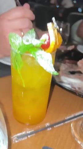 The video shows a close-up view of several drinks on a wooden table. In the foreground, a tall glass filled with an orange-colored drink, ice, mint leaves, and a slice of orange with a cherry is visible. A straw lies on the table in front of the glass. To the right, a martini glass holds a similar orange-colored drink topped with what appears to be passionfruit pulp. In the background, other drinks and people are out of focus. The overall setting appears to be a restaurant or bar.