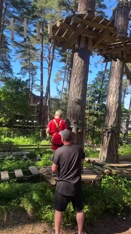 The video captures a sunny day at a treetop adventure park in Jūrmala, Latvia. The scene is dominated by towering pine trees, some of which have been incorporated into the park's obstacle courses. Wooden platforms and bridges are suspended at various heights between the trees, creating a challenging and exciting environment.

In the foreground, a man wearing a black t-shirt and shorts stands on a wooden platform, looking up at a young boy. The boy, dressed in a red t-shirt and a safety harness, is perched on the shoulder of the man, seemingly observing the adventure park. Another man, wearing a black cap, is also visible, positioned behind the boy. Further in the background, other individuals can be seen navigating the course, adding to the sense of activity and fun. The presence of the adventure park, with its wooden structures and lush greenery, suggests a recreational setting, likely a tourist attraction. The bright sunlight and clear blue sky indicate pleasant weather conditions, perfect for outdoor activities. The overall mood appears to be one of excitement and familial engagement, with the man providing a vantage point for the boy to take in the surroundings.
