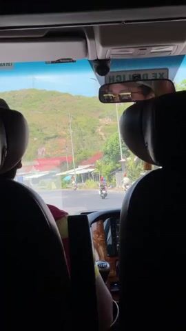 This media captures a view from inside a vehicle, likely a van, looking out through the windshield. The scene depicts a sunny day in Chí Thạnh, Vietnam, with lush green hills in the background. A rural road runs through the middle ground, lined with houses that have red tile roofs and some smaller buildings. Several motorbikes are visible on the road, with at least three riders clearly identifiable. One person is wearing a red helmet, another a blue shirt, and a third appears to be wearing a white shirt.

In the foreground, we see the backs of two headrests and the interior of the vehicle, including a gear shifter and part of a dashboard with a screen. A rearview mirror reflects the faces of two passengers wearing sunglasses, suggesting a bright, possibly warm, day. The overall mood is casual and observant, capturing a glimpse of everyday life in a scenic Vietnamese landscape.

Visible text includes "XE DU LỊCH" on a sticker in the upper right portion of the windshield, which translates to "Tour Vehicle" or "Tourist Car" in Vietnamese. There is also partial text on a sticker in the upper left, "NGƯỜI LÁI XE...", which is likely part of "NGƯỜI LÁI XE TỐT", meaning "Good Driver".
