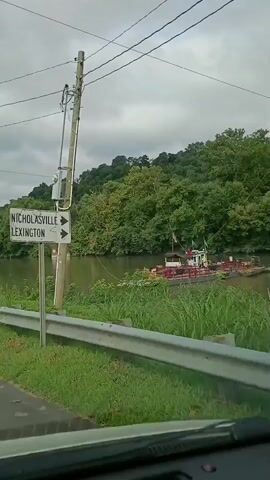 The video captures a view from inside a vehicle looking out at a rural roadside. A signpost with directional arrows pointing to "NICHOLASVILLE" and "LEXINGTON" stands prominently on the left. Beyond the sign, a wide river flows, with a barge carrying cargo and a small cabin visible on its deck. The riverbanks are lined with lush green trees and tall grass. On the right side of the road, a series of traffic signals and signs are mounted on a pole, including a red traffic light, a diamond-shaped "FERRY" sign, and a rectangular sign indicating "100 FEET." A red octagonal "STOP" sign is also present. A white Tesla SUV is seen driving away from the camera in the second half of the video. The sky is overcast, suggesting a cloudy day. The road surface appears to be asphalt, and a metal guardrail runs along the edge of the road. The overall scene depicts a ferry crossing area in a natural, wooded environment.