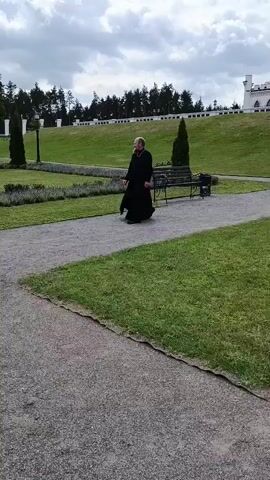 A person wearing a long black robe walks down a path in a park. In the background, a white castle with pink accents sits atop a grassy hill. Several other people, including a man and woman in casual attire and a child, walk towards the castle stairs. The sky is cloudy.
