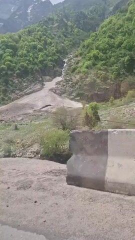 The camera is inside a vehicle, likely a car, traveling along a paved road. The view is primarily of a mountain slope on the right-hand side of the road. The mountain is covered in green trees and rocky areas. A large expanse of light-colored material, possibly a mudslide or debris flow, stretches down the slope. The mountain has a few patches of snow at its summit, suggesting high altitude or cooler temperatures. Concrete barriers are present along the side of the road. There's a utility pole to the right. The sky is overcast. The sound indicates music playing.