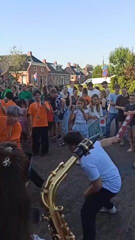 The video shows an outdoor event with a crowd of people, likely a street festival or parade, in Warffum, Netherlands. The scene is set on a paved street lined with brick houses. The time of day appears to be late afternoon or early evening, with warm lighting and clear skies.

The main subjects are two young individuals, one wearing an orange t-shirt and the other a light blue t-shirt, engaged in a playful "sword fight" using cylindrical, red and white objects that resemble foam pool noodles. Both are energetically moving and posturing.

Surrounding them is a diverse crowd of spectators of all ages, many of whom are watching the activity. Some people are holding phones to record. In the background, Dutch flags are visible on the houses, confirming the location. The atmosphere seems festive and joyful, with people of different ages enjoying themselves.

There is some visible text on the red and white "swords" but it is too blurry to read clearly. The overall impression is of a lively community gathering with a lighthearted performance.