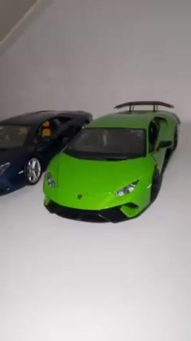 The video shows two toy Lamborghini cars, one blue and one green, sitting side-by-side on a white surface. The blue car is a Lamborghini Aventador, and the green car is a Lamborghini Huracan. The camera slowly zooms in on the cars, showing their details. The blue Aventador has a yellow interior, while the green Huracan has a black interior and a large rear wing. The background is white, and the lighting is soft.