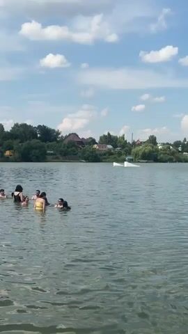The video shows a lively scene at a beach resort on a sunny day with scattered clouds. The first clip captures people swimming and playing in the water of a lake or pond. Several individuals are visible, some with inflatable toys, enjoying the refreshing water. The background shows a wooded shoreline with houses peeking through the trees.

The subsequent clips transition to the sandy beach area. People are seen relaxing on lounge chairs under umbrellas, sunbathing, and strolling along the shoreline. Children are playing on the sand, with one young child crawling near the water's edge. Beach umbrellas and lounge chairs are set up in neat rows, indicating a well-maintained recreational area. The overall atmosphere appears to be one of leisure and enjoyment on a summer day.