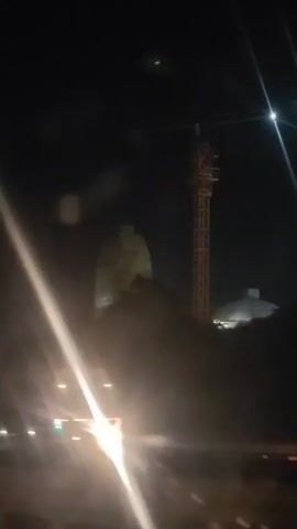 A nighttime video shows a large, illuminated, dome-shaped structure, possibly under construction, with scaffolding and a crane visible. The structure is dark green and appears to be made of stone or concrete. There are lights illuminating the base of the structure. The crane is tall and yellow, with construction materials visible at the top. The video is shaky, suggesting it was filmed from a moving vehicle. The background is dark, with some distant lights and trees visible.