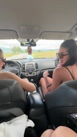 Two women are inside a car. The woman on the left is in the driver's seat. She has long brown hair in a ponytail, is wearing sunglasses and a black tank top, and is tapping on the center console with her left hand. The woman on the right is in the passenger's seat. She has long dark hair, is wearing black-rimmed glasses and a black top, and is clapping her hands together. She is facing the driver, with her right leg bent at the knee and her left leg resting on the dashboard. The car is moving on a paved road with fields and trees on either side. There are hills in the distance. The sky is overcast.