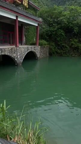 The video shows a serene landscape featuring a river and traditional Chinese architecture. The river's water is a distinctive shade of green. Lush greenery covers the surrounding hills and structures.  A stone bridge supports a building with a red-trimmed facade.  As the camera pans, a long white building with a red base and black roof tiles is revealed, situated along the riverbank. A metal rope barrier comes into view, suggesting a pathway for visitors.