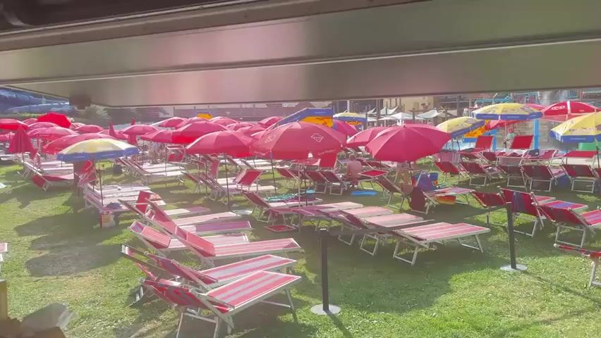 The video shows a park-like area with many empty lounge chairs and red umbrellas set up on green grass. In the background, there are water slides and people swimming in a pool. There is a serving counter in the foreground with some boxes and a tray of food. The sun is shining and it appears to be a warm day. The video seems to be taken from under an awning or roof.