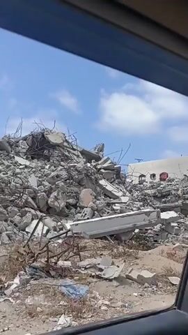 The video shows a view from inside a vehicle looking out at a pile of rubble. The rubble consists of concrete debris, rebar, and other building materials. Behind the rubble, a damaged building with white walls and windows is visible. The sky is blue with some clouds. The ground is covered in dirt and dry vegetation. The footage appears to be taken during daylight. There are no people visible in the video.