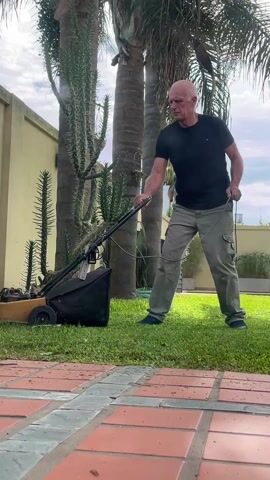 A bald man in a black shirt and tan cargo pants stands on a green lawn next to a red-brick patio, using a yellow electric lawnmower with a black collection bag. He pulls the cord to start the mower, but it doesn't start.  The lawn is bordered by a light-yellow wall. In the background there are tall green cacti and palm trees. The sky is partly cloudy.