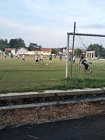 The video shows a soccer game being played on a grassy field. There are several players in white and black uniforms and others in blue and white uniforms, spread across the field. One player with the number 50 on their back is walking toward the center of the field. The camera is positioned behind a soccer goal, offering a view of the field from that perspective. In the background, there are trees and buildings, suggesting the field is located in a residential area. The sky is overcast.