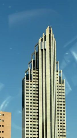 The video shows a tall, modern skyscraper with a stepped, pointed roof against a clear blue sky. The building is mostly white with vertical dark accents and numerous rectangular windows. There are metal structures protruding from the upper section of the building. To the left of the skyscraper, a portion of a shorter, tan building is visible. Towards the bottom of the frame, palm trees and a black structure, possibly a fence or barrier, are present. The camera appears to be moving, as evidenced by the slight blurring and changing perspective. The overall scene depicts an urban environment on a sunny day.