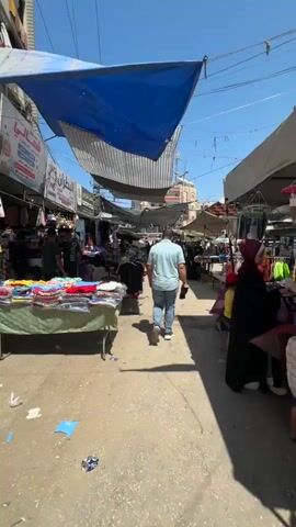 The video opens showing a wide shot of a marketplace with stalls set up on either side of a path. Several awnings made of fabric are stretched overhead to provide shade. A man in a light blue shirt and jeans walks away from the camera down the center of the path. More people are visible further down the path, browsing at stalls or walking. On the left side of the path, a stall displays colorful clothing on tables and racks. On the right side, other stalls also display merchandise. The ground is paved and appears somewhat worn. The sky is clear and blue. As the video progresses, the camera follows the man in the blue shirt. Another man carrying a plastic water jug and a bag walks towards the camera from further down the path. The camera continues to move forward, capturing more of the market activity and the variety of goods on display.