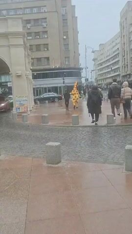 The video shows a city scene in rainy weather with several pedestrians walking on a wet sidewalk. 

In the distance, a person dressed in a bright yellow giraffe costume walks among the other pedestrians. Buildings of varying heights line both sides of the street, and a black car is parked near the curb. The pedestrian area is marked by short concrete posts. The ground is wet from the rain, creating reflections of the surrounding environment.