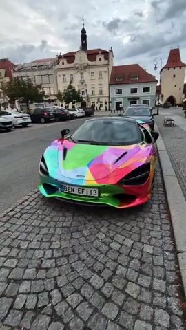 The video shows a McLaren 720S sports car parked on a cobblestone street. The car is wrapped in a vibrant, multi-colored, abstract design. The surroundings include other parked cars and buildings with traditional architecture. The sky is overcast.