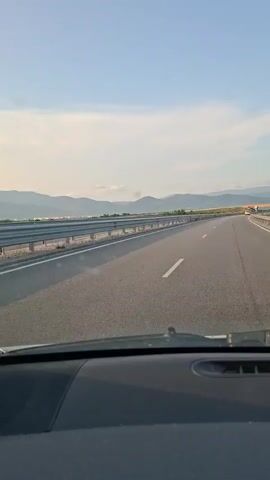 The video shows a point-of-view shot from inside a vehicle driving on an empty highway. The road ahead is clear, with mountains visible in the distance under a clear sky with a few clouds. The sun appears to be setting, casting a warm glow on the landscape. The vehicle's dashboard is partially visible in the foreground.