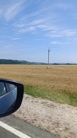 The video is taken from the passenger seat of a moving car, looking out the window. The scene is a rural landscape on a clear day with a bright blue sky and scattered white clouds. A vast field of dry, golden grass or wheat stretches across the middle ground, with a line of green grass along the edge of the road in the foreground. In the distance, rolling hills are visible under the horizon. Several utility poles with wires are spaced throughout the field. A side mirror of the car is visible on the left side of the frame. The car appears to be traveling on a paved or gravel road.