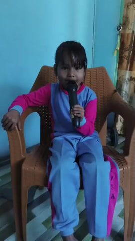 A young girl wearing a pink and blue tracksuit is sitting on a plastic chair and holding a black microphone to her mouth. She is looking directly at the camera. The background is a light blue wall with a patterned curtain on the right.